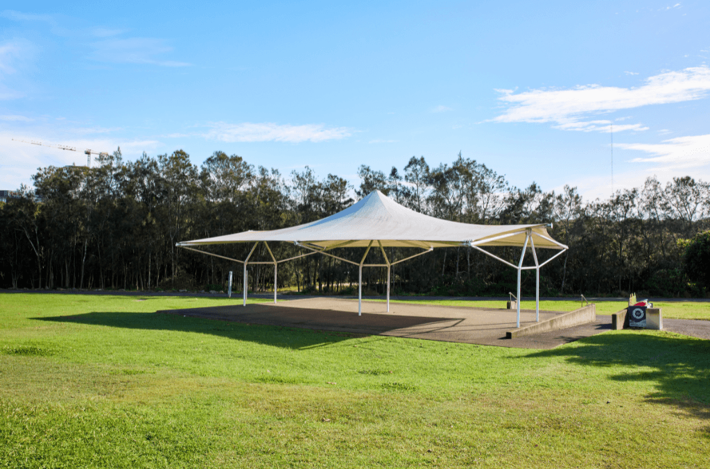 A white shade sail structure covers an open paved area in a grassy park, with trees lining the background under a clear blue sky.